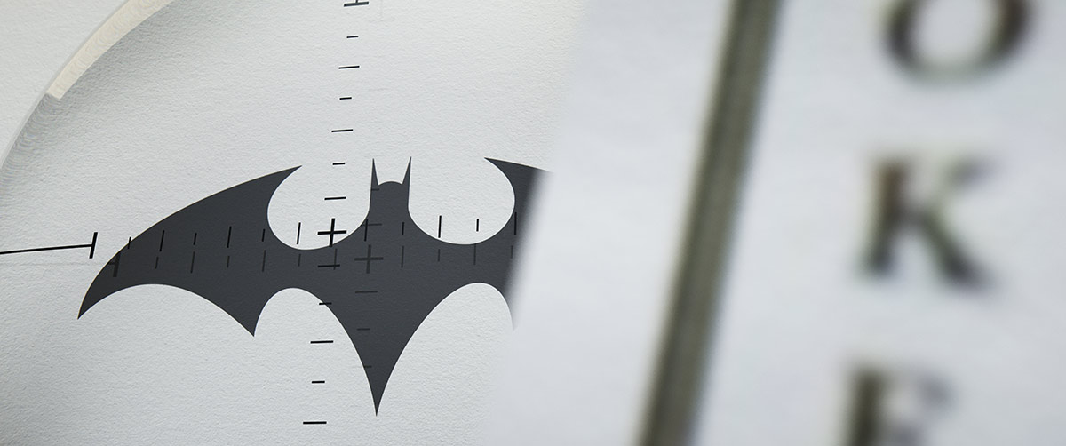 A close-up look-development frame focusing on a small bat logo viewed through a circular glass lens with crosshair reticle telemetry, positioned next to a blurred playing card.