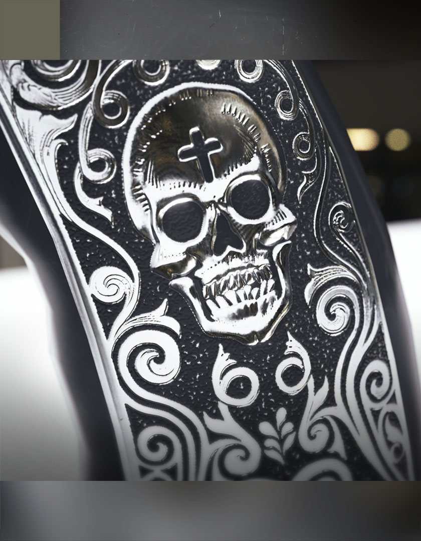 An extreme macro crop focusing on a custom deep-etched steel gun receiver component, highlighting a stylized skeletal face graphic nestled among intricate baroque swirl scrollwork.