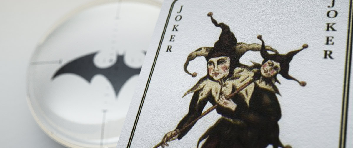 A graphic style frame pairing an illustrated Joker playing card on the right with a circular lens scope overlay showing a small bat logo and technical targeting crosshairs on the left.