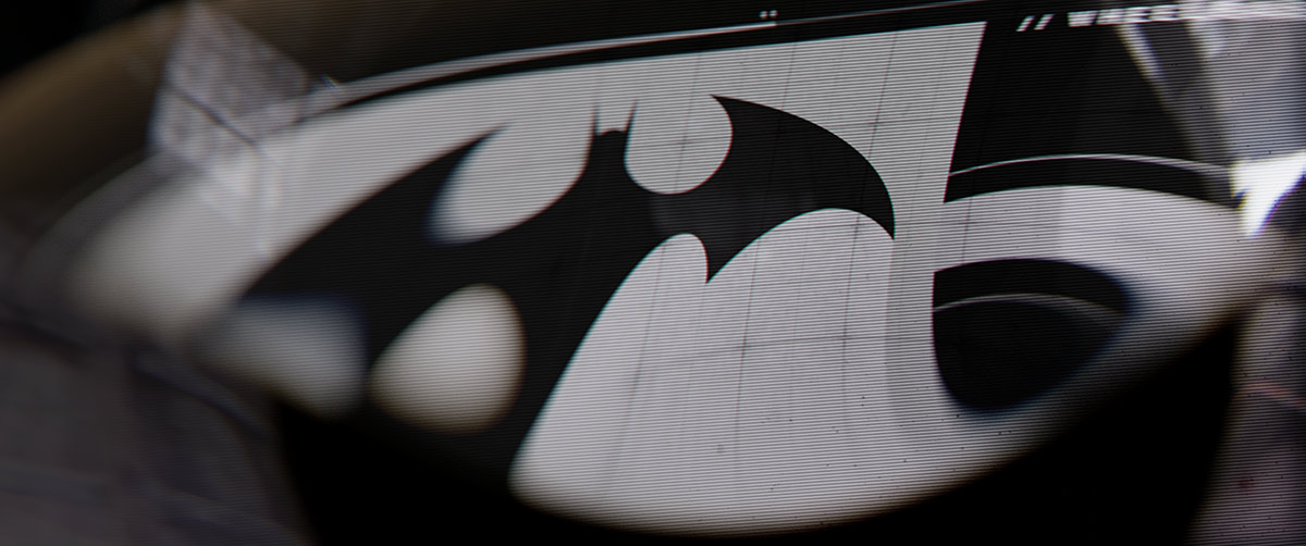 A close-up style frame showing the redesigned bat logo emblem displayed on a curved monitor interface with digital scanlines and edge distortion.