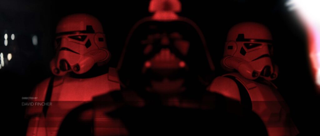 A dramatic cinematic style frame showing a reverse rack focus shift where the two flanking 3D Stormtrooper models are rendered in sharp optical clarity, while the central silhouette of Darth Vader is completely blurred into the dark background under intense red studio lighting.