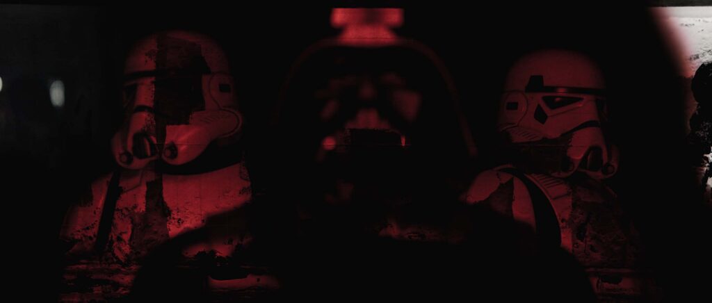 A dark, cinematic style frame showing the blurred central silhouette of Darth Vader flanked by two out-of-focus Stormtroopers under deep red lighting, uniquely overlaid with heavy, organic grunge textures and simulated ink splatters across the white armor.