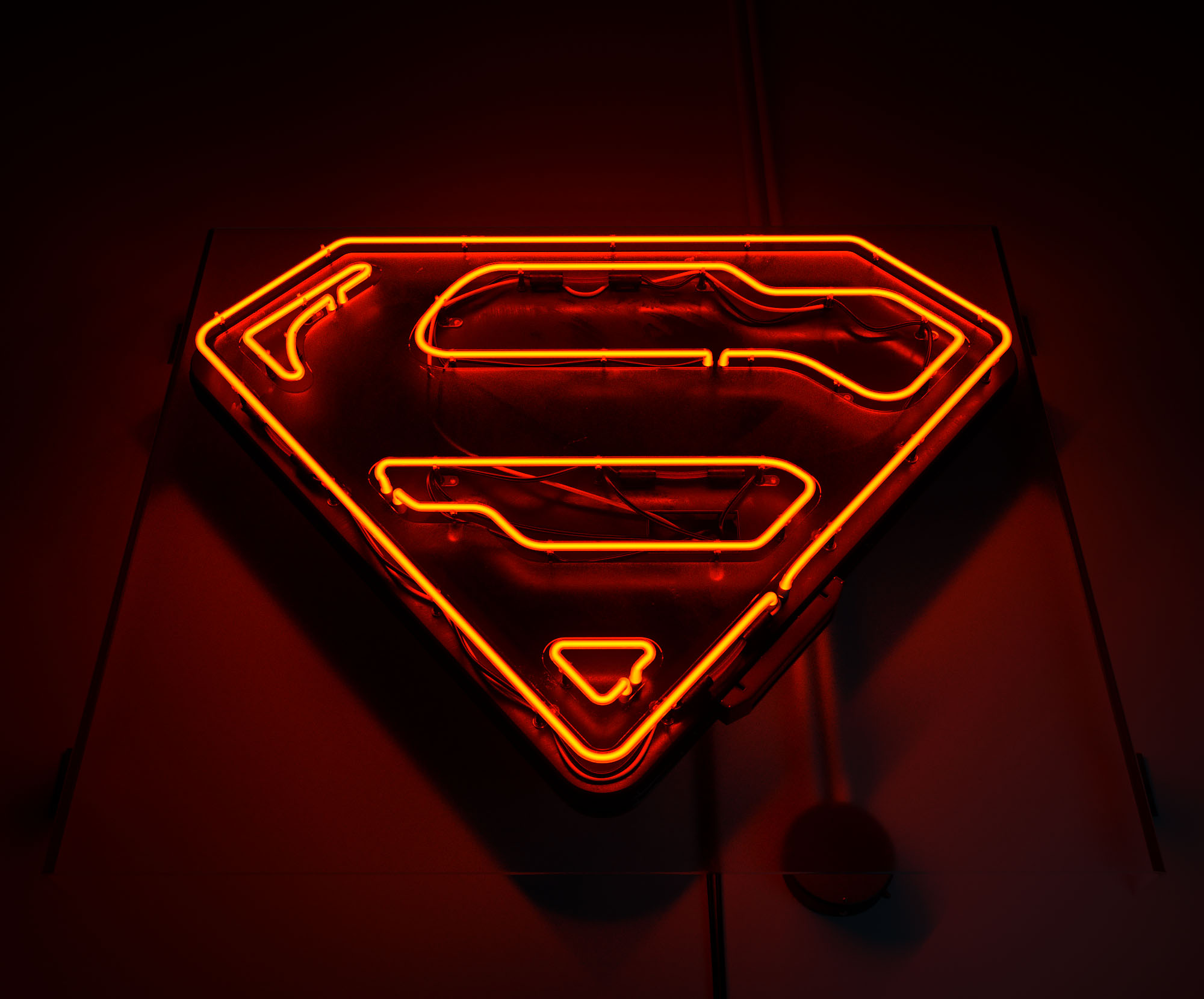 A 3D render of the contemporary Superman shield emblem stylised as an active, glowing red-orange neon sign set within a dark environment, casting a warm volumetric light across the glass tubing, metallic backing structure, and internal hardware wiring.